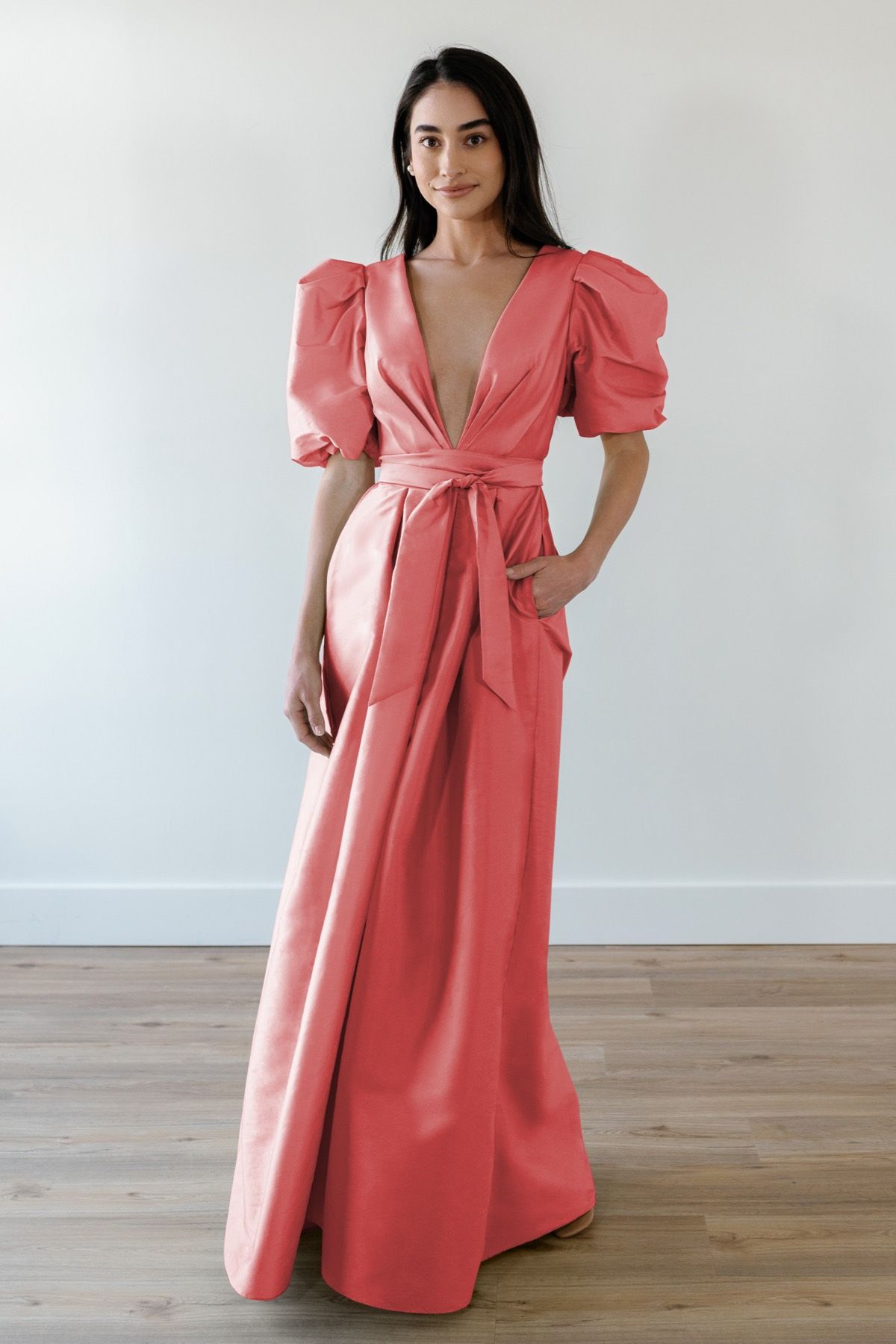 A woman in a long pink dress with puffed sleeves is standing on a wooden floor.