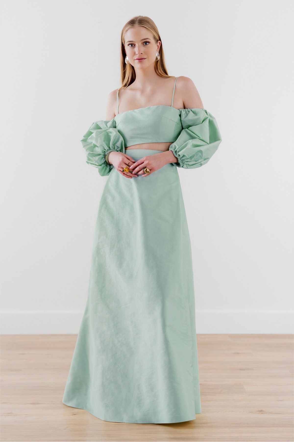 A woman in a long green dress with puffed sleeves is standing on a wooden floor.