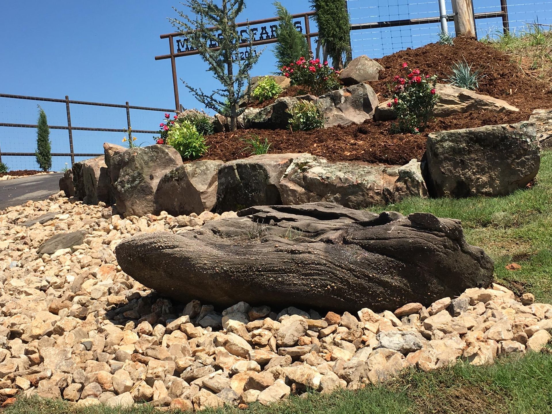 Rock sculpture in a landscaped garden with pebbles, flowers, and a wooden fence under a blue sky
