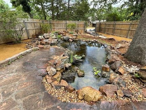 Backyard pond with rocks, wooden fences, and a large tree beside a stone patio
