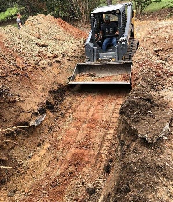 Excavator grading a narrow muddy trench on a dirt path in a wooded area