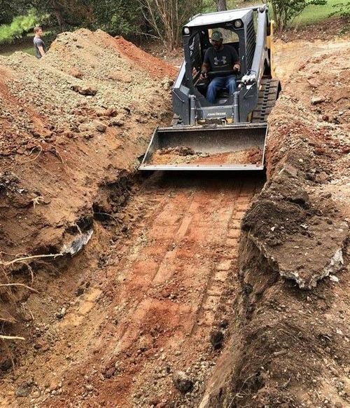 Excavator grading a narrow muddy trench on a dirt path in a wooded area