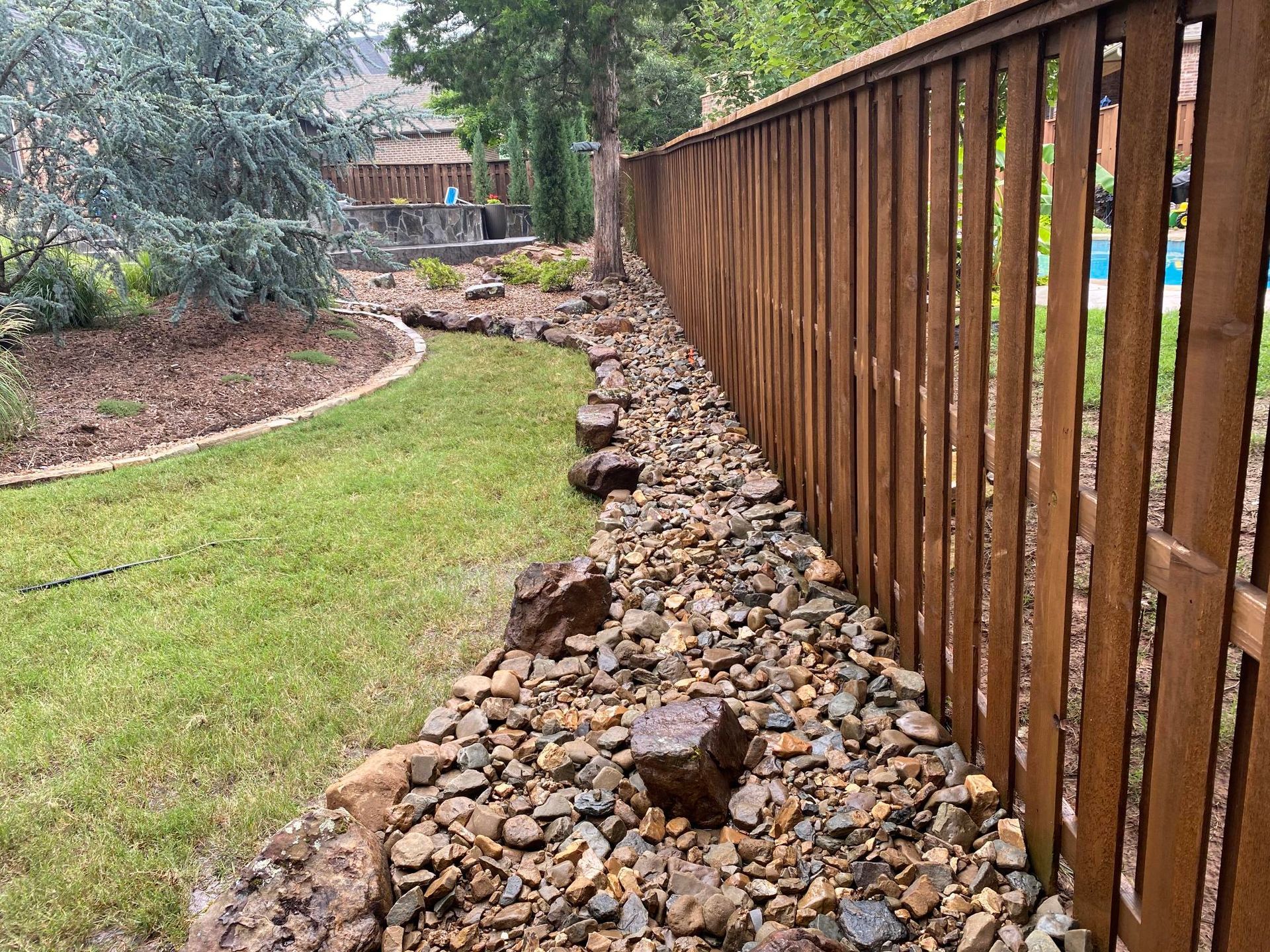Rock-lined drainage beside a wooden fence, with a curved lawn and garden in the background.