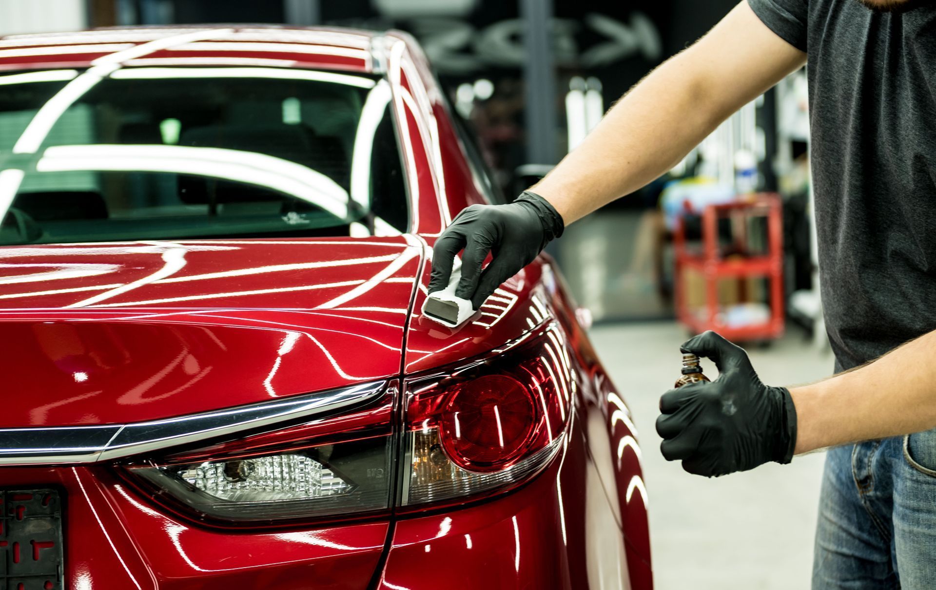 Person applying sealant to a red car's taillight, wearing black gloves.