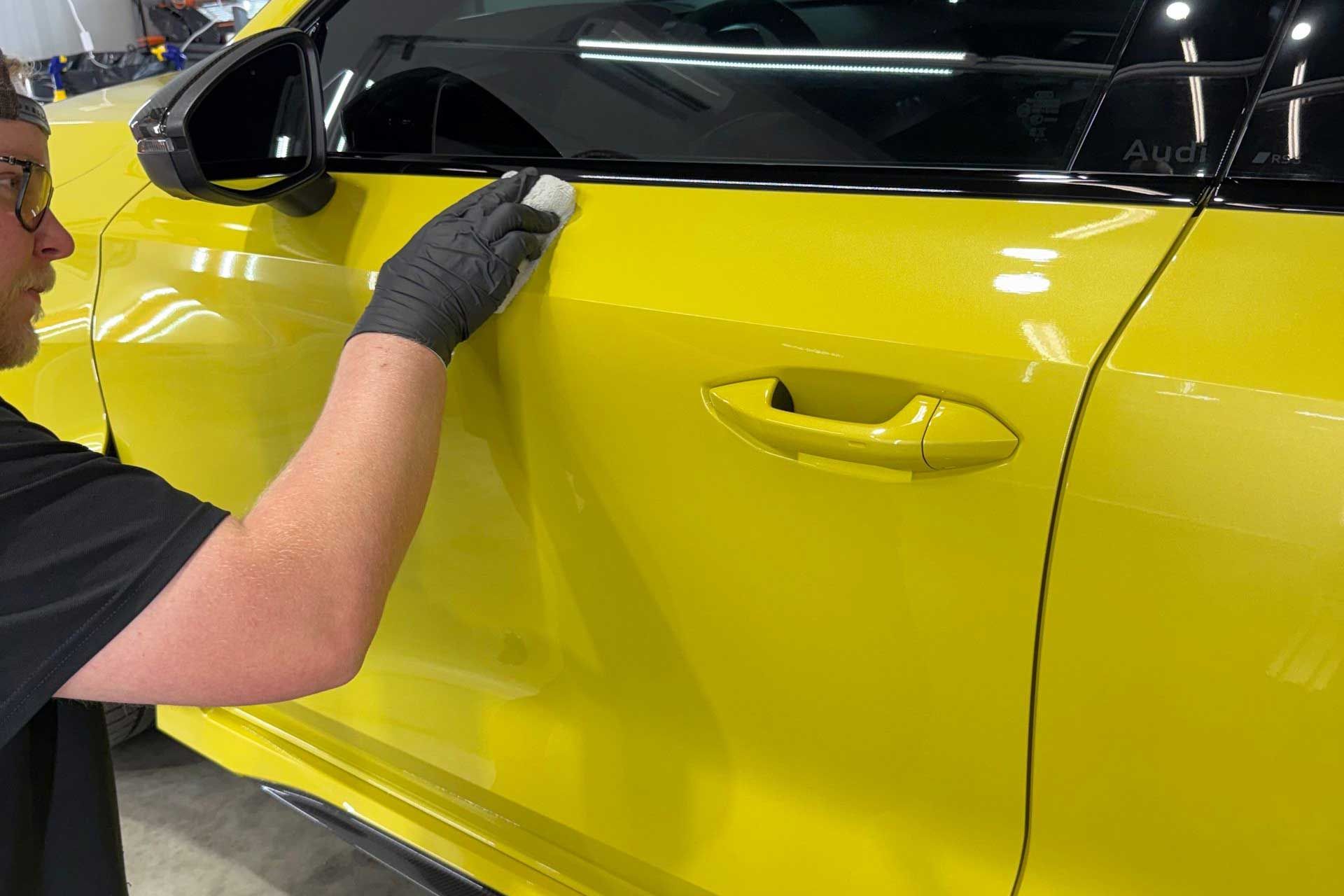 A person wearing black gloves applies a coating to a yellow car door.