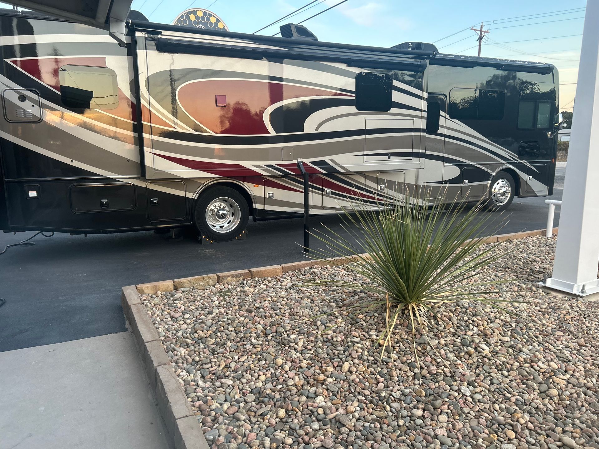 Large RV parked in front of a building with gravel landscaping. Brown, red, and gray exterior.