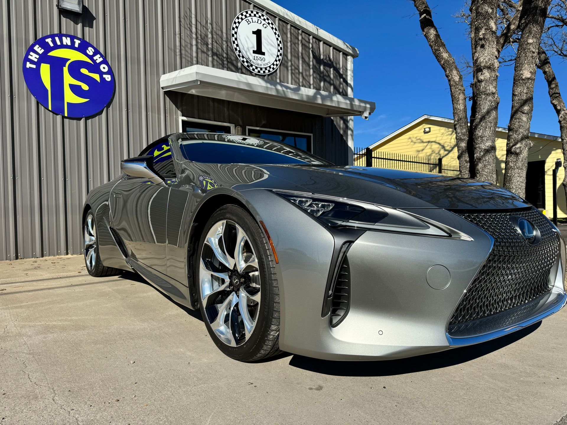 Silver Lexus coupe parked in front of a shop with a blue and yellow sign; sunny day.