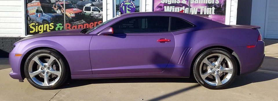 Purple Camaro parked outside a shop advertising signs, banners, and window tint.