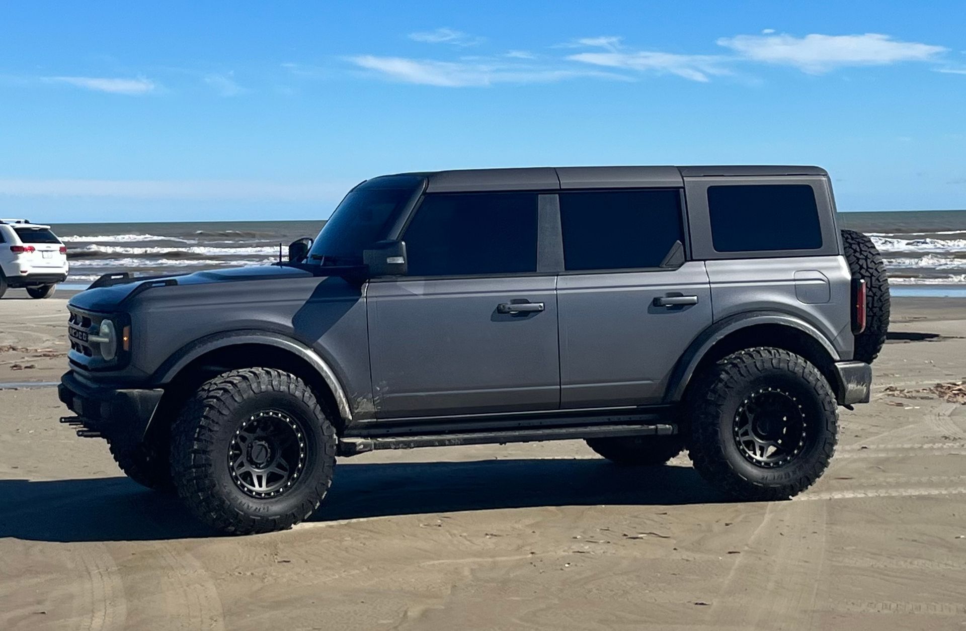 Gray Ford Bronco on a sandy beach with dark tinted windows and black wheels. Ocean in background.