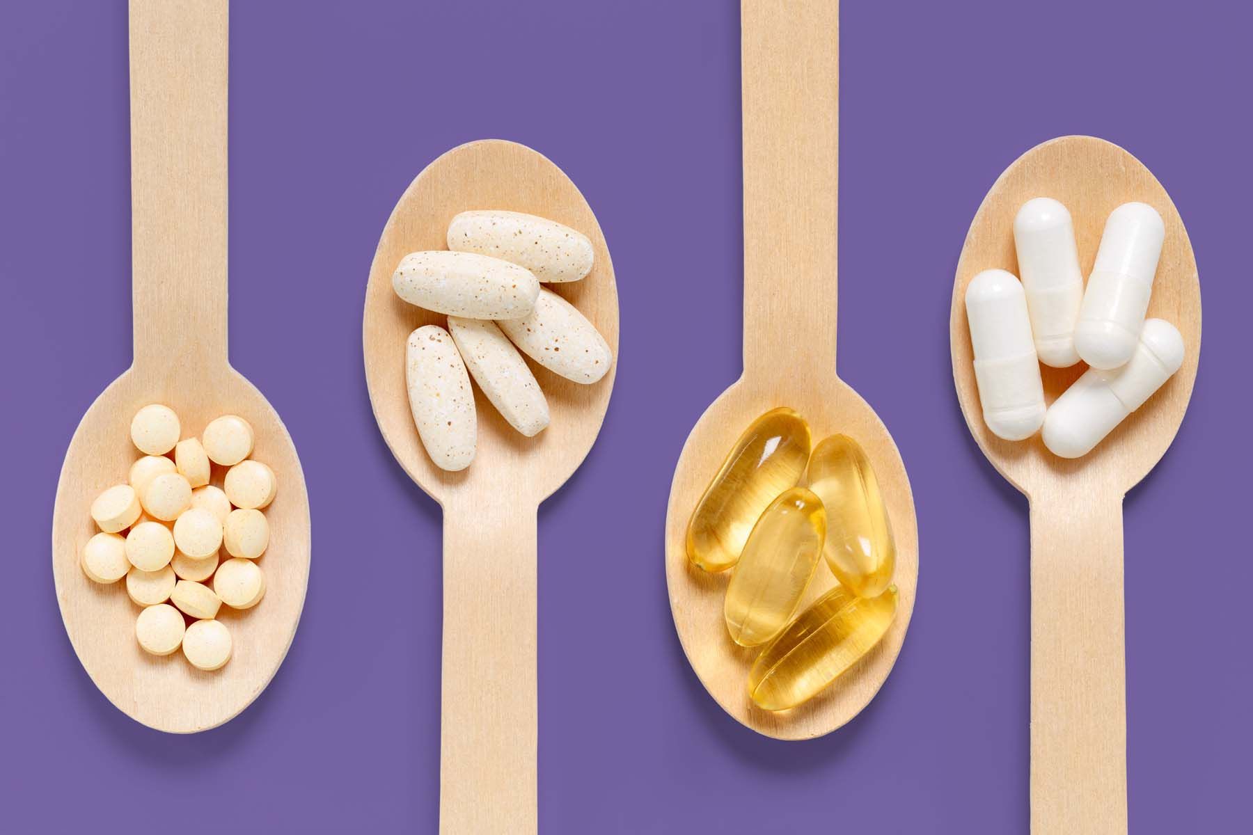 Four wooden spoons filled with different types of pills on a purple background.