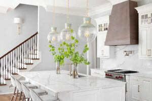 A kitchen with white cabinets , a large island , a stove , and a staircase.