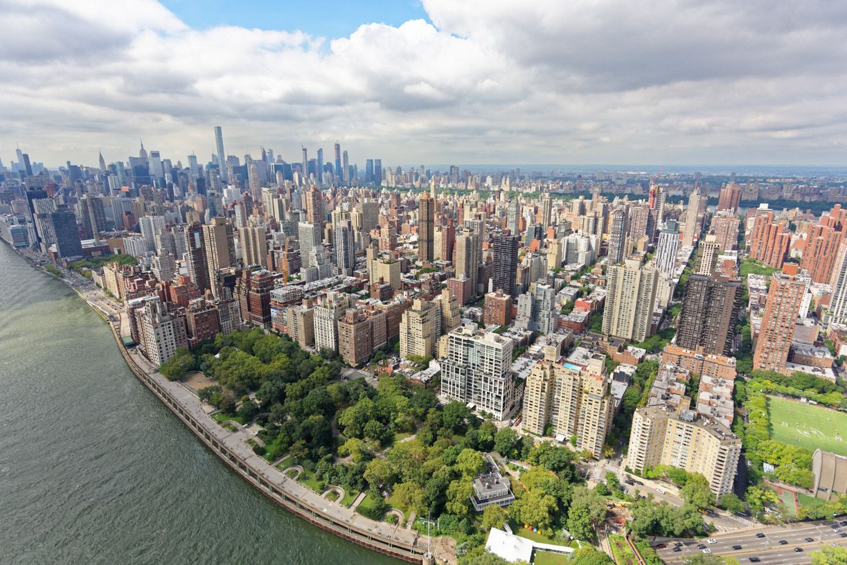 Aerial view of a dense city skyline along a waterfront, with a park and cloudy sky.