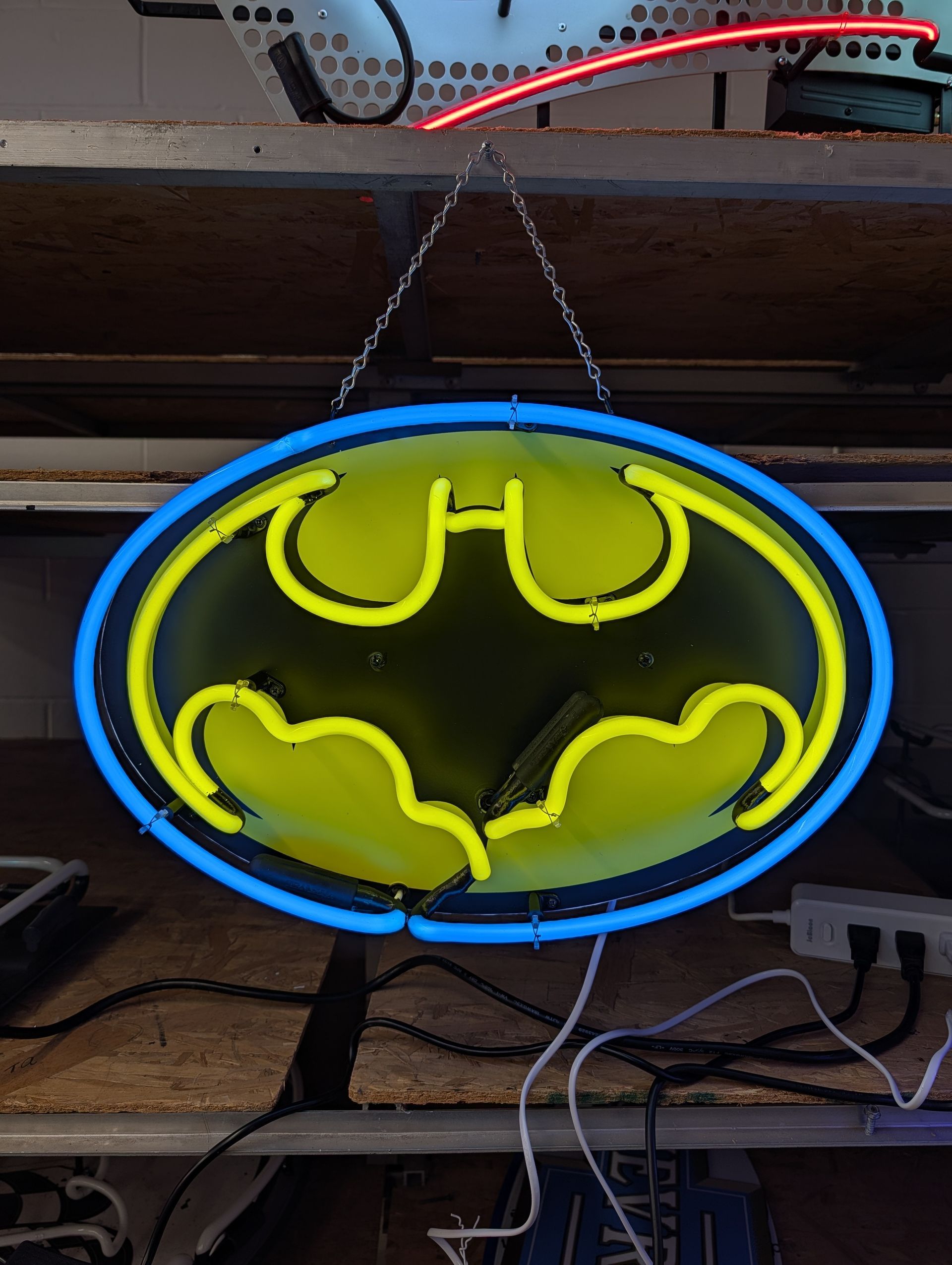 Batman logo neon sign glowing yellow and blue, hanging indoors above a shelf.