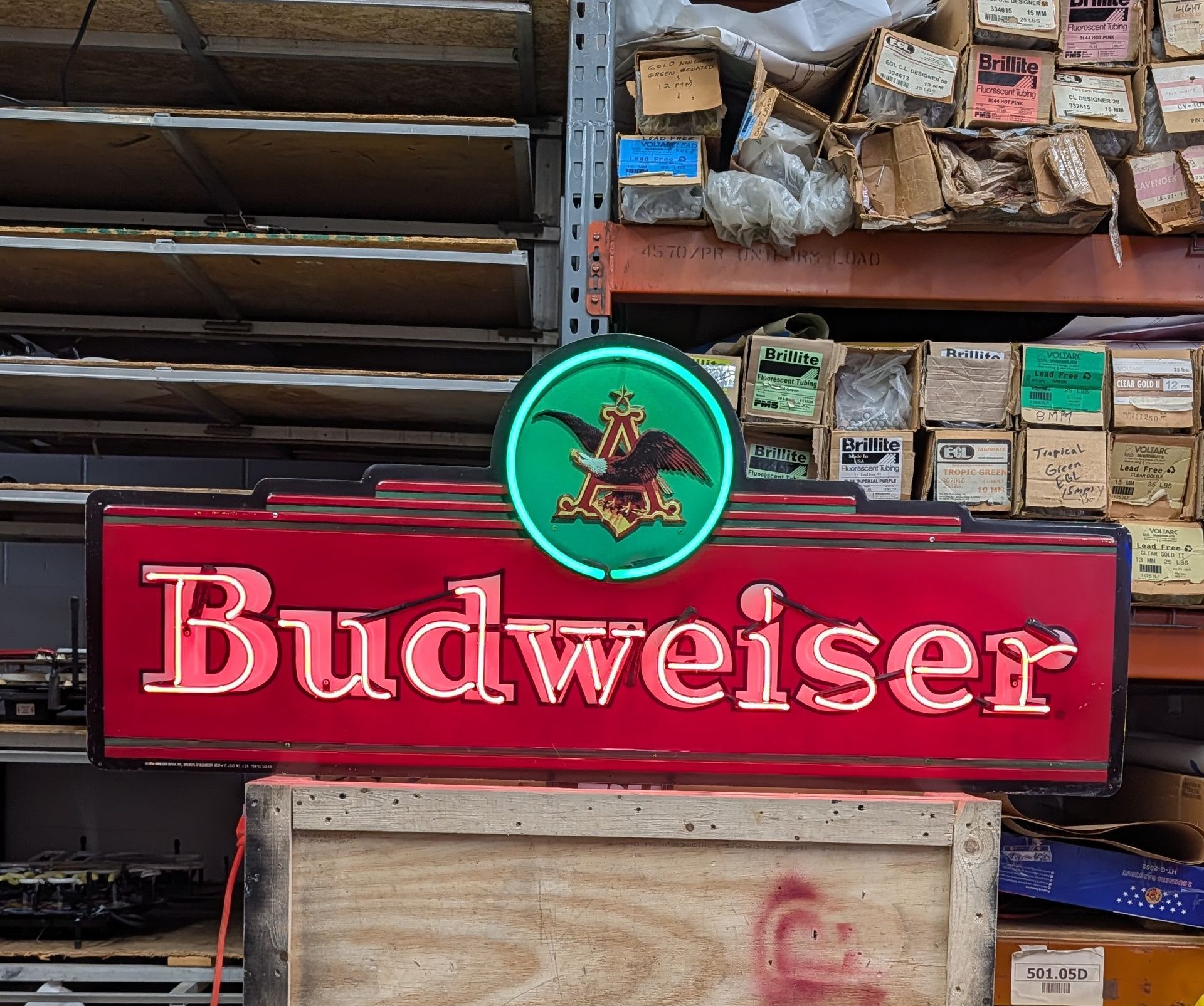 Red Budweiser sign with green circular logo on a wall of stacked crates and boxes.