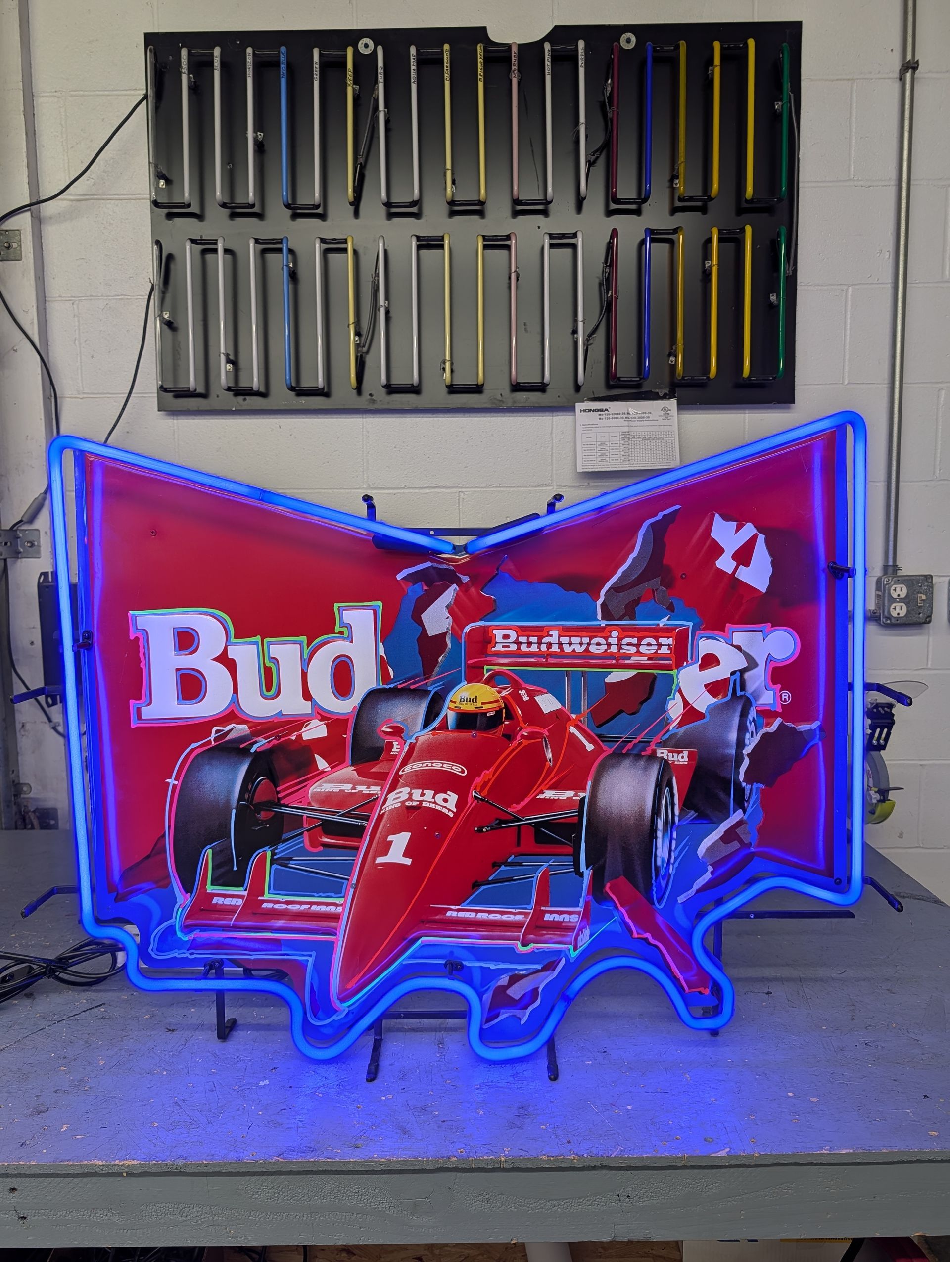 Red neon Budweiser sign with a red Formula 1 car in front, displayed in a workshop.