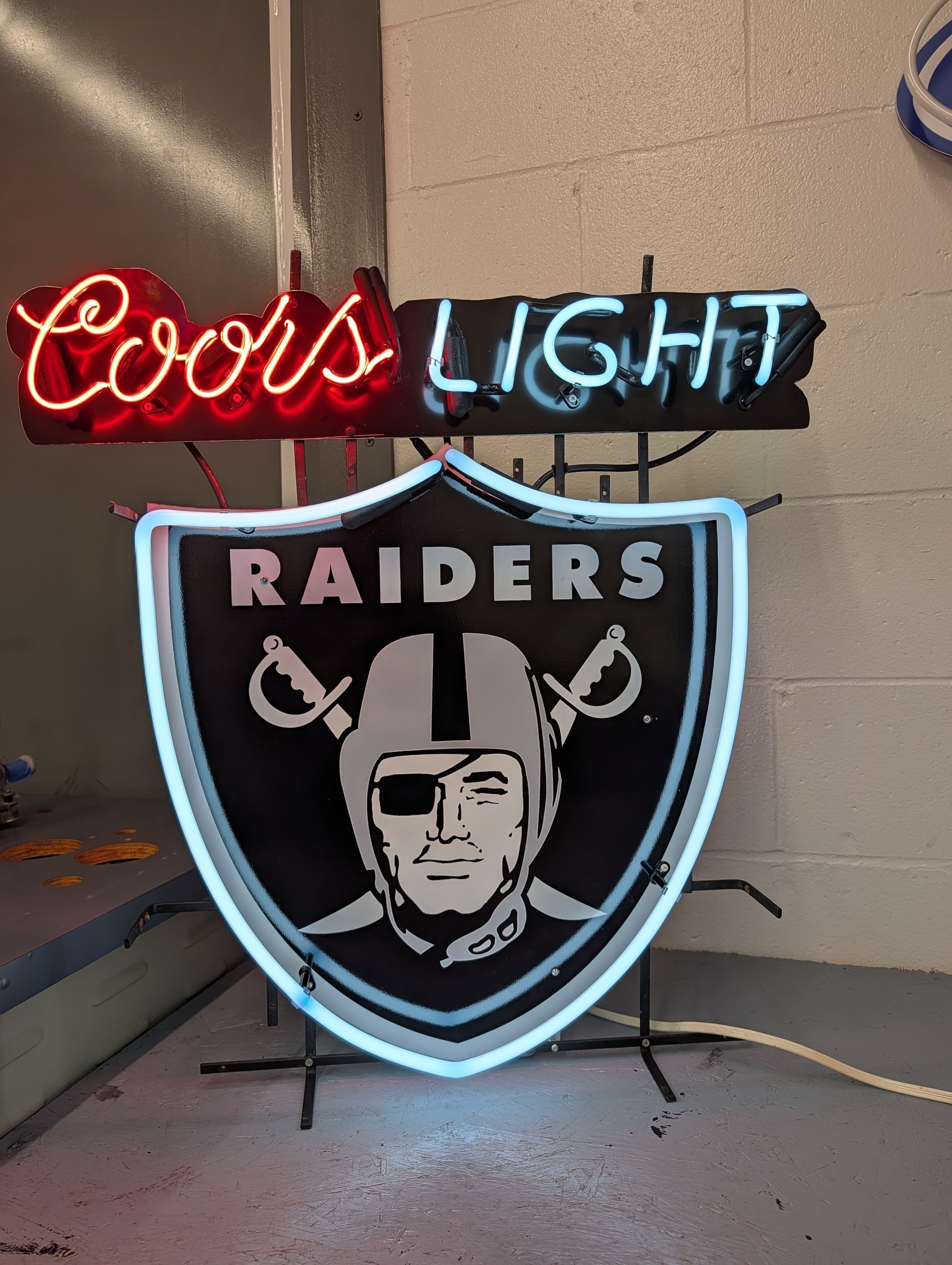 Coors Light neon sign above a Raiders shield logo neon sign on a wall-mounted stand