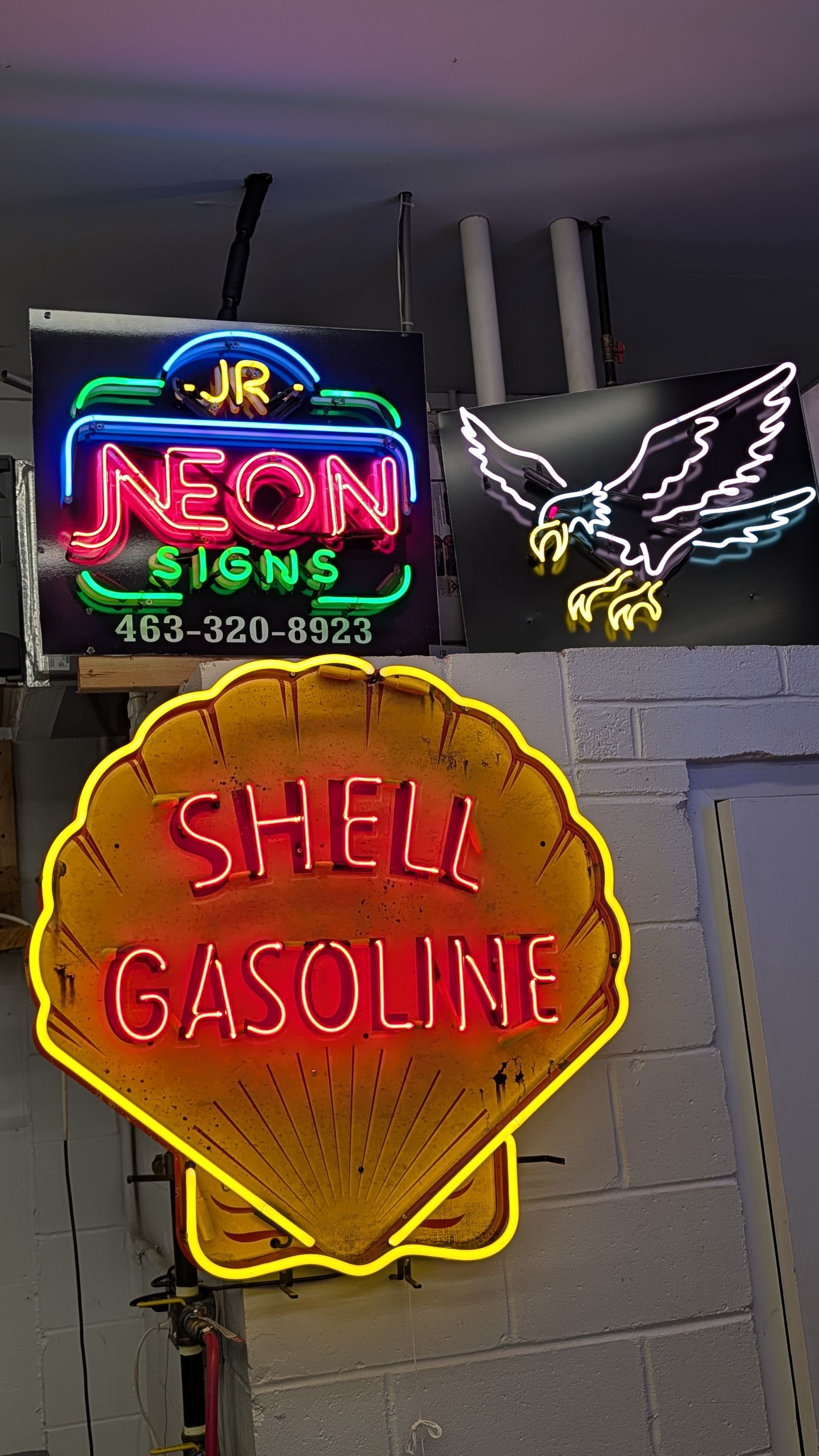Neon signs on a wall, including a Shell Gasoline sign and a neon eagle logo above it.