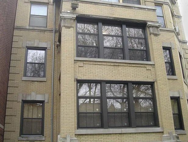 Three-story tan brick apartment building with large dark-framed windows and bare trees reflected in glass