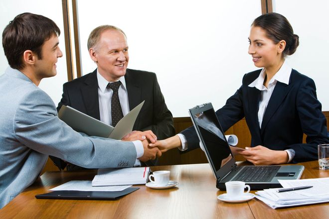 Three professionals shaking hands across a conference table with laptops and documents in an office