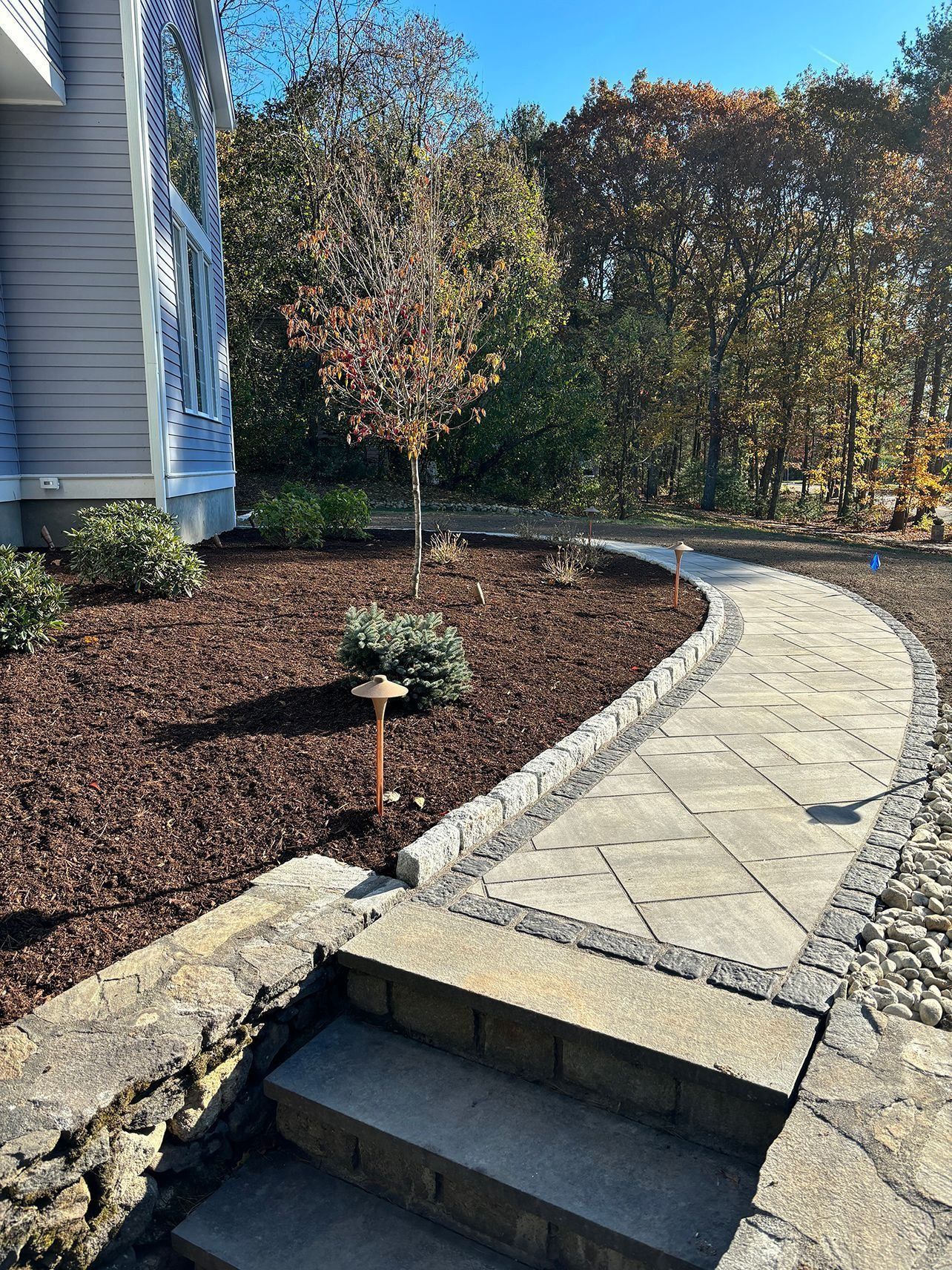 Stone pathway leading to a house with steps. Brown mulch, small tree, and fall foliage in background.