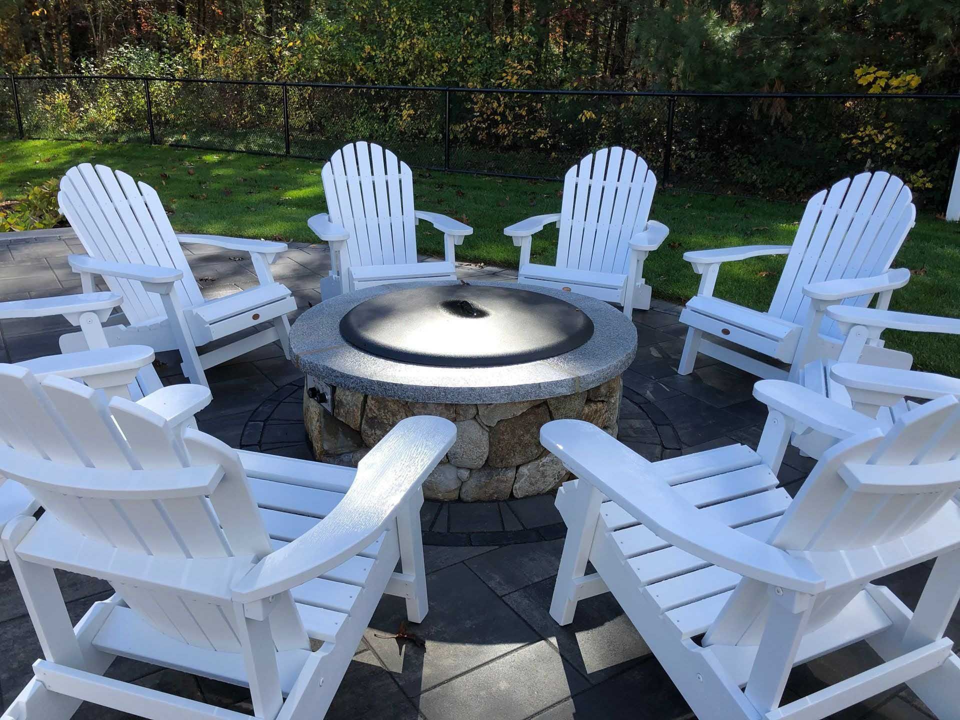 White Adirondack chairs circle a stone fire pit on a patio.