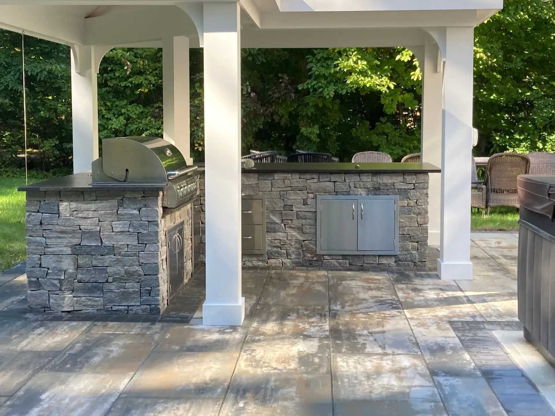 Outdoor kitchen with stone facade, built-in grill, and stainless steel doors, under a white pergola.