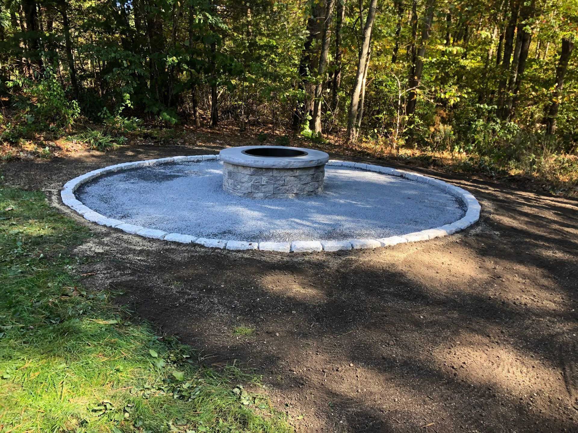 Fire pit made of stone in a gravel circle with a stone border, set in a wooded area.
