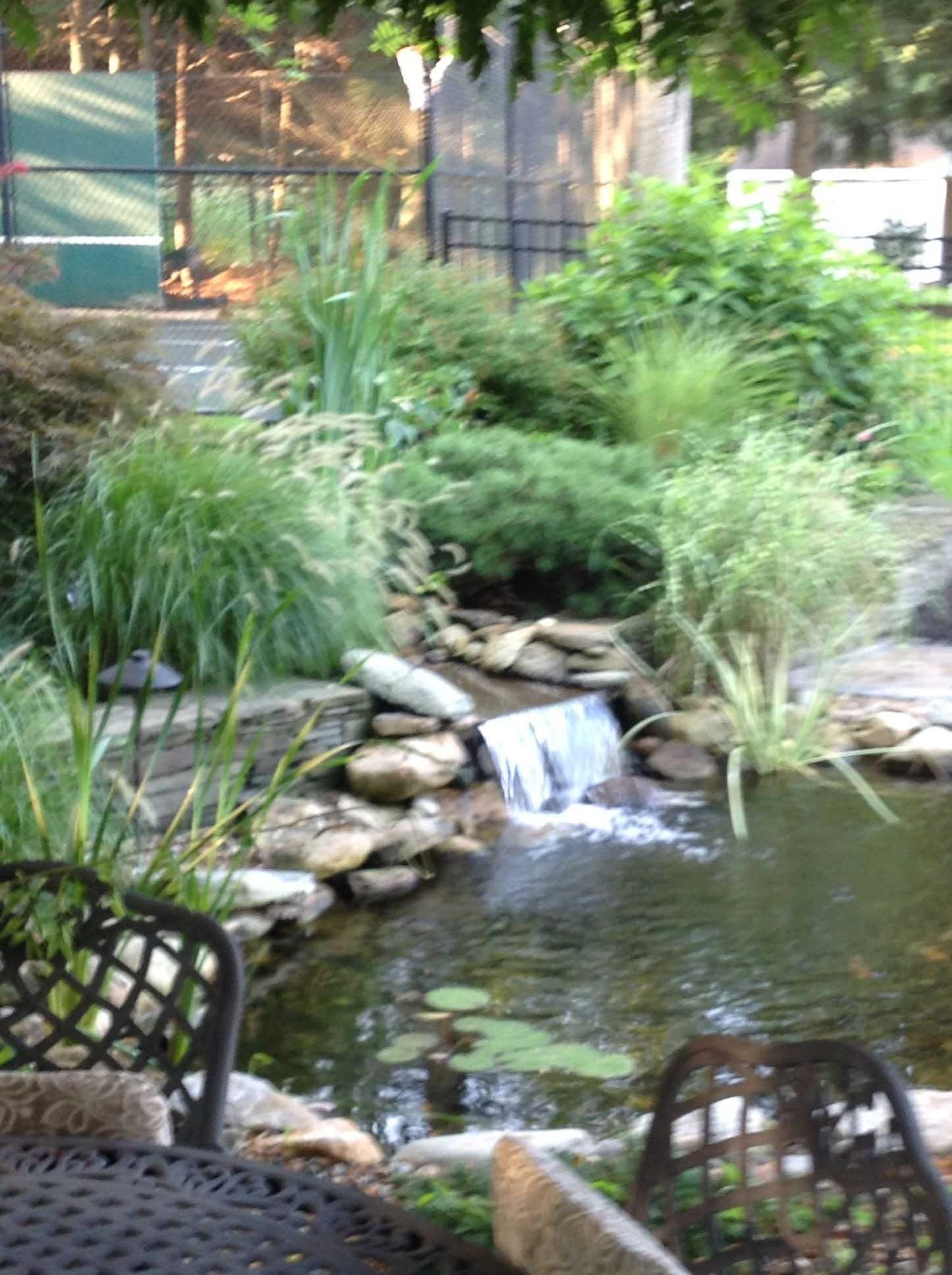 A small waterfall cascades into a pond surrounded by lush greenery. Two chairs and a table are in the foreground.