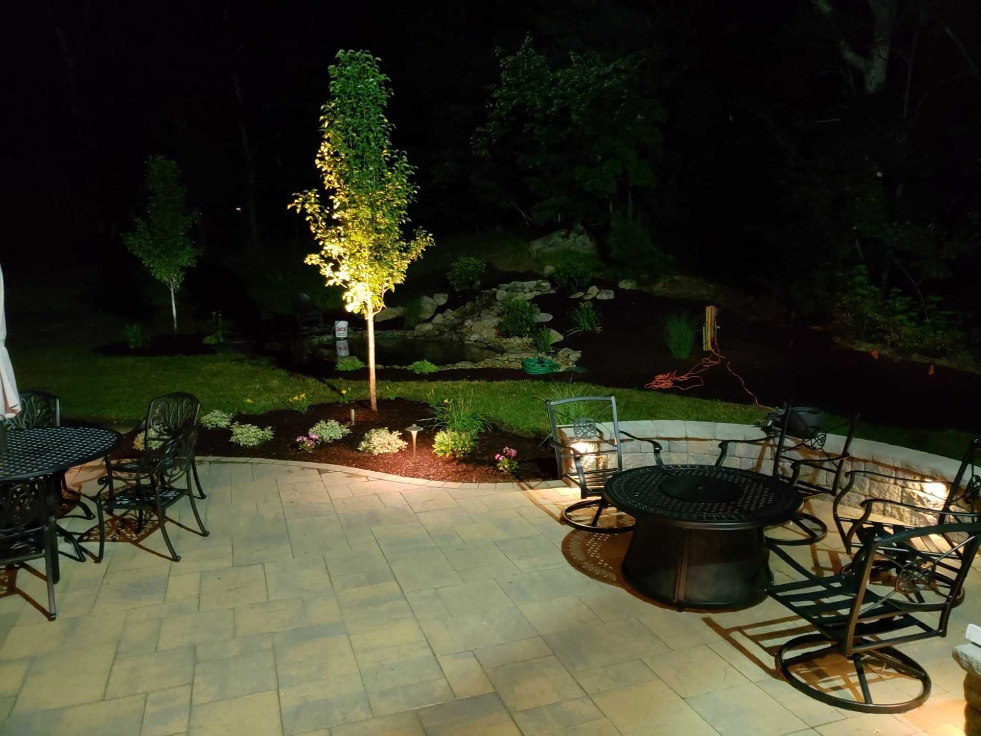 Patio with dark wrought iron furniture, uplighting illuminating a tree and landscaping at night.