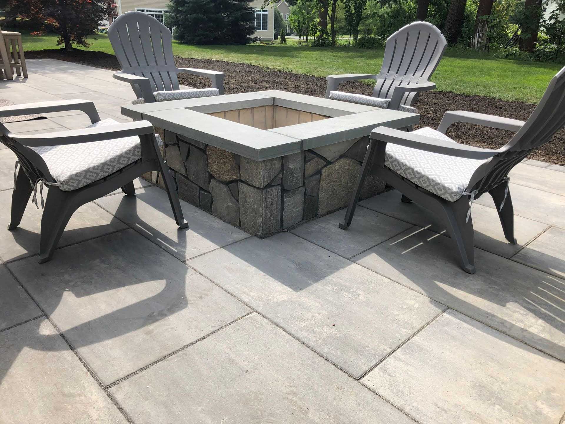 Four gray chairs surround a stone fire pit on a paved patio.