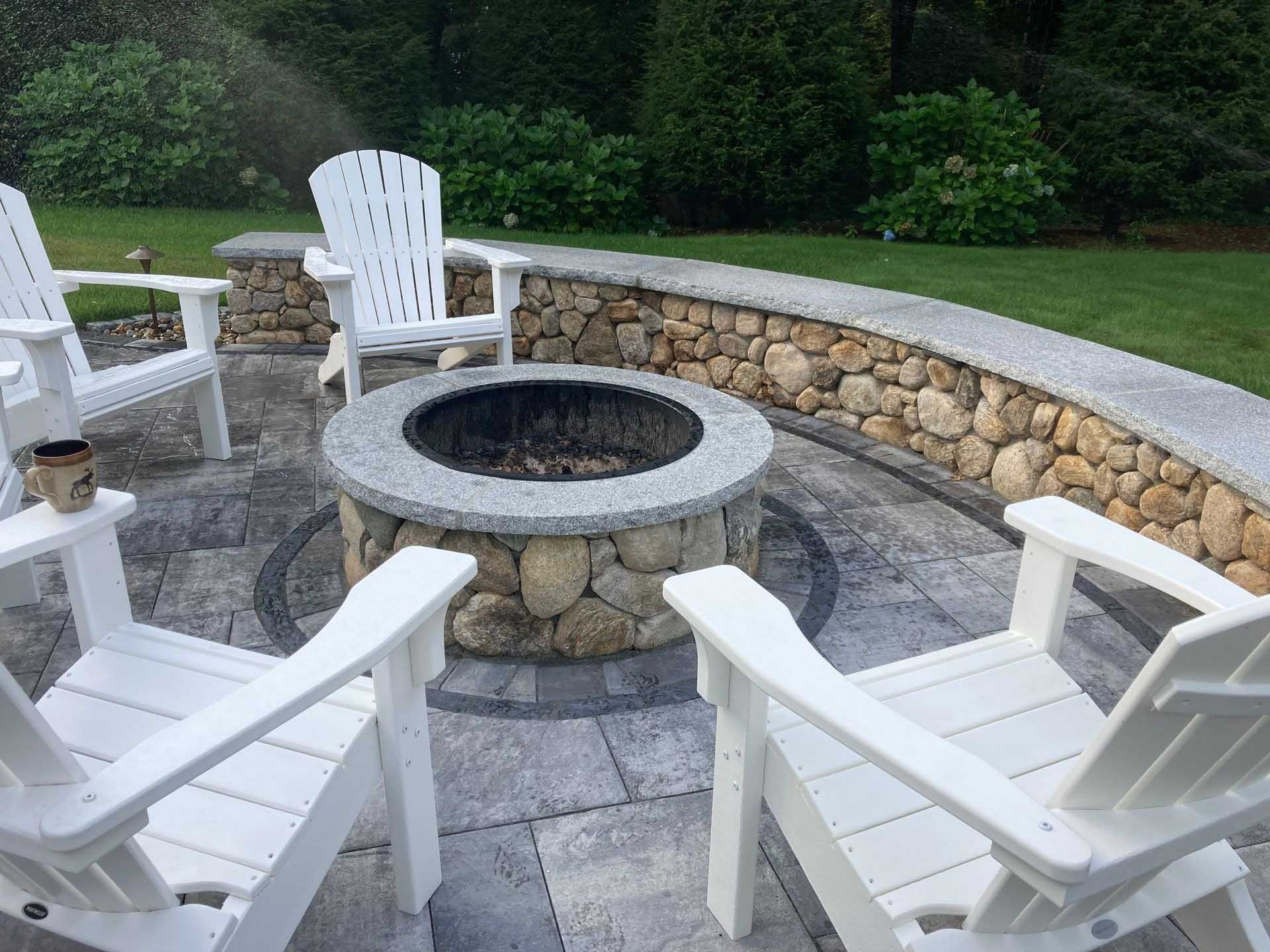 Fire pit with Adirondack chairs on a stone patio, surrounded by a low stone wall and greenery.
