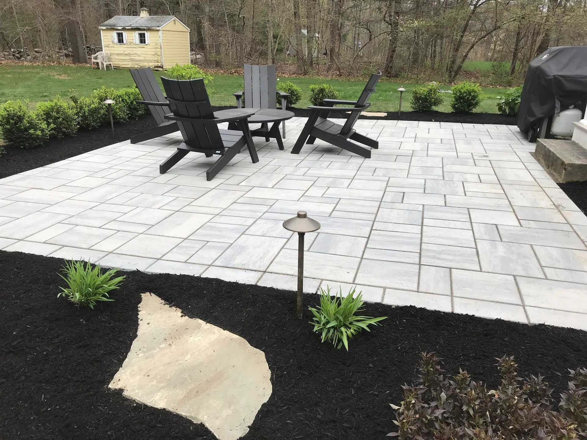 Patio with black chairs, gray pavers, and dark mulch, surrounded by green bushes, with a shed in the background.