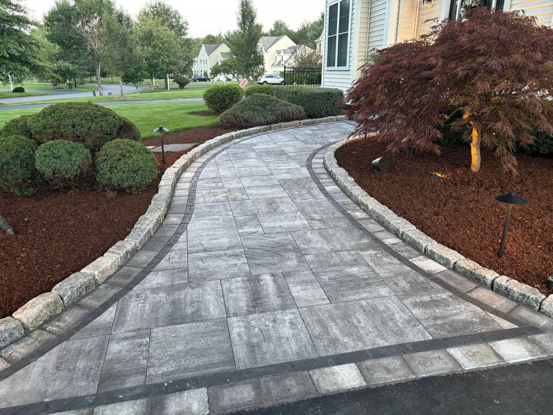 Stone walkway curves toward a house, edged with mulch, plantings, and a small tree.
