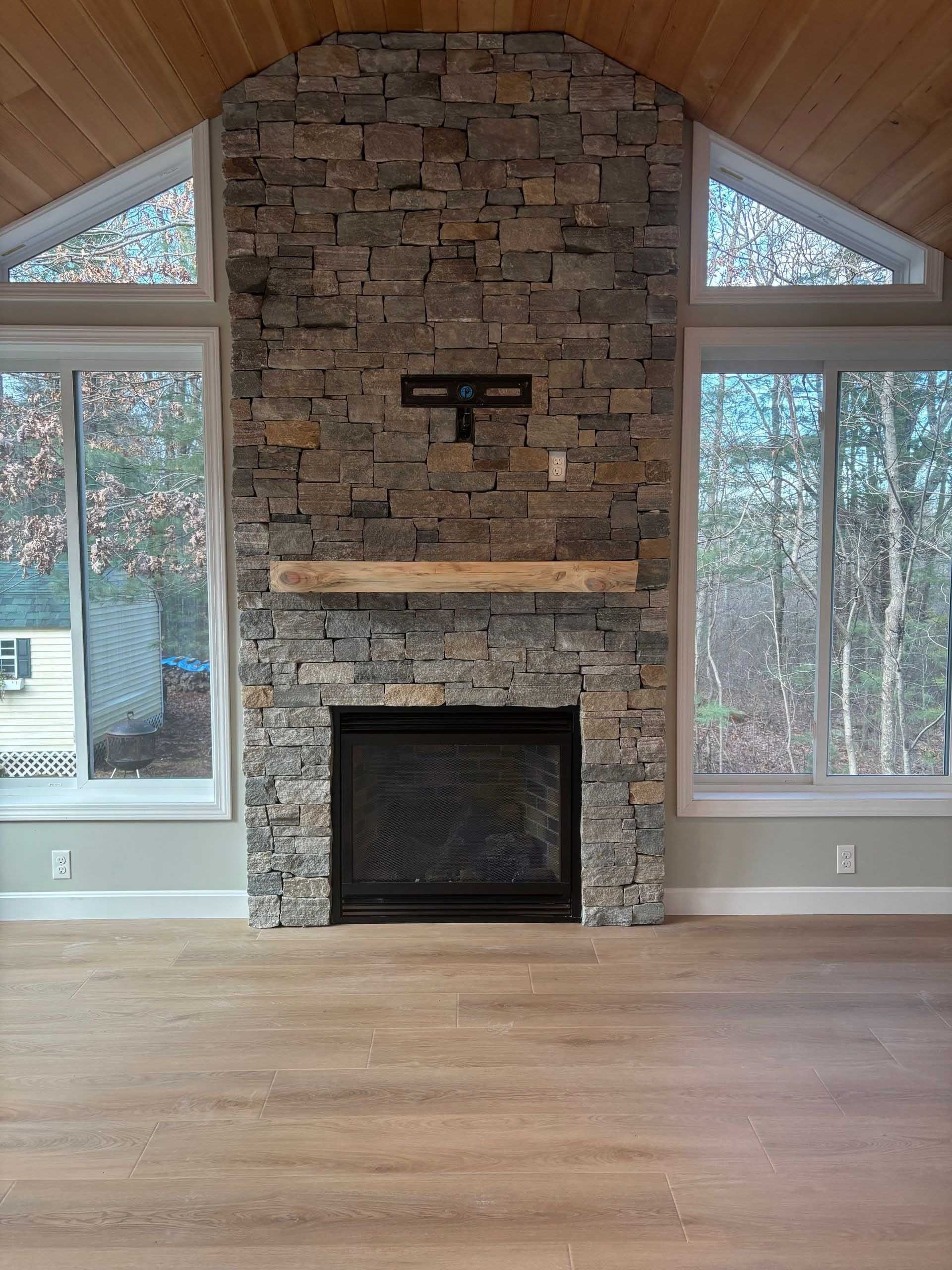 Fireplace with stone facade, wooden mantel, and windows; natural light on wood floor.