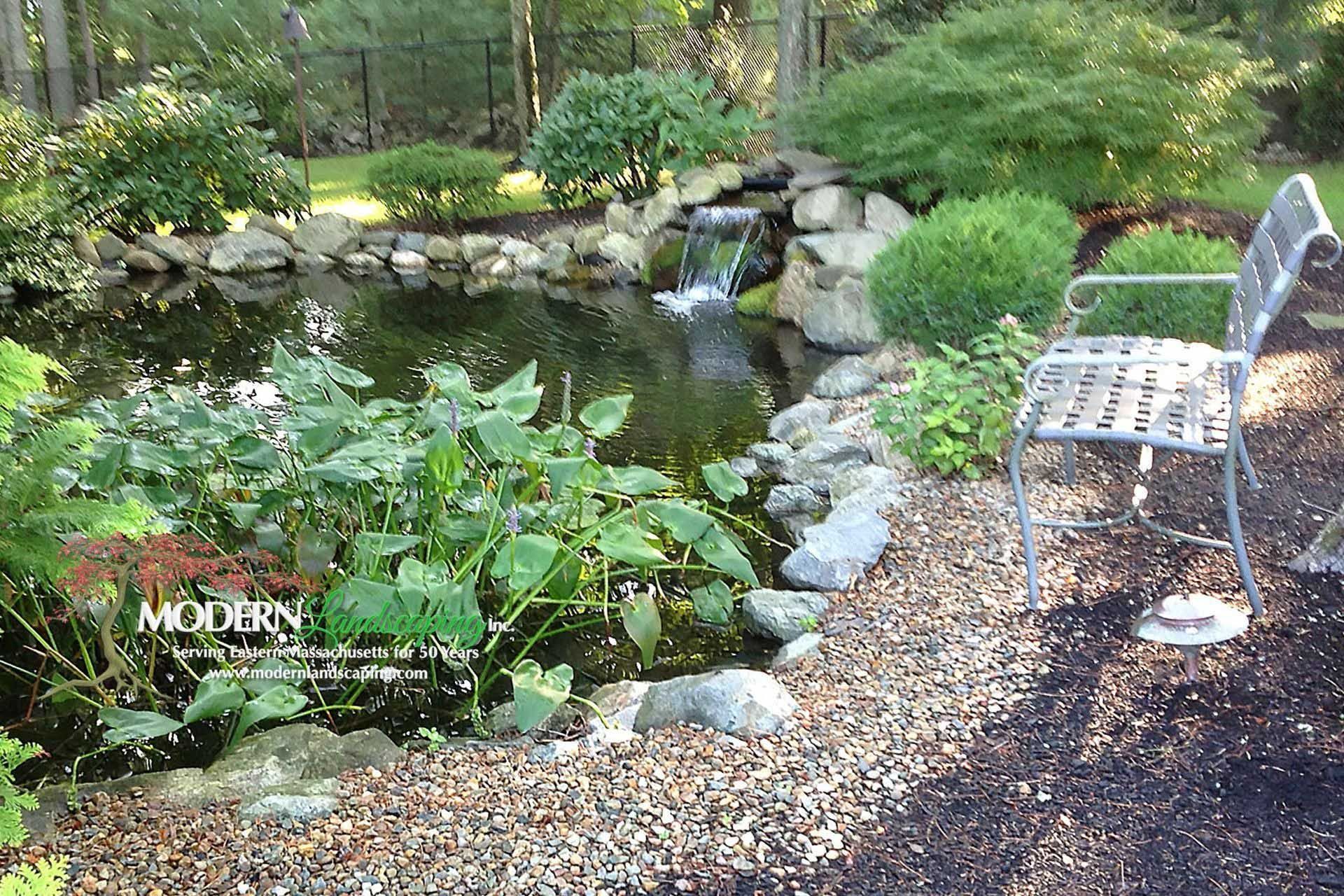 Pond with waterfall, lush greenery, and a metal chair on a gravel path.