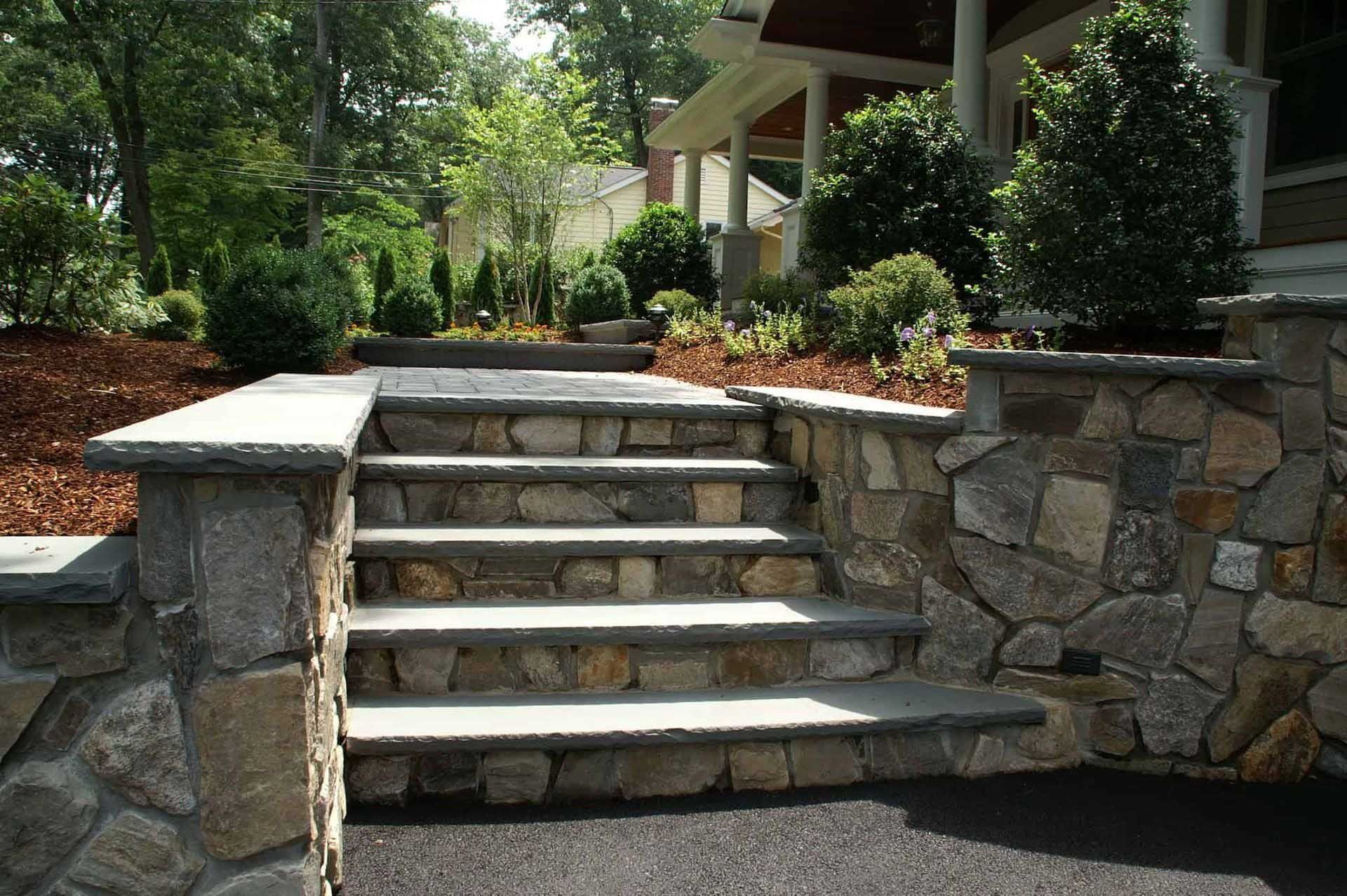 Stone steps leading up to a house, flanked by stone walls and landscaping.