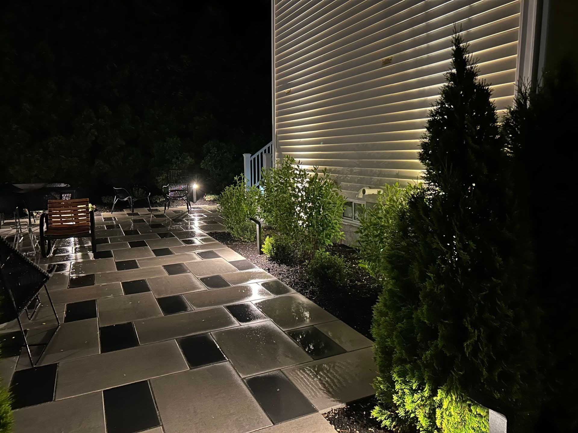 Nighttime view of a patio with lit paving stones, illuminated building and foliage, and a bench.