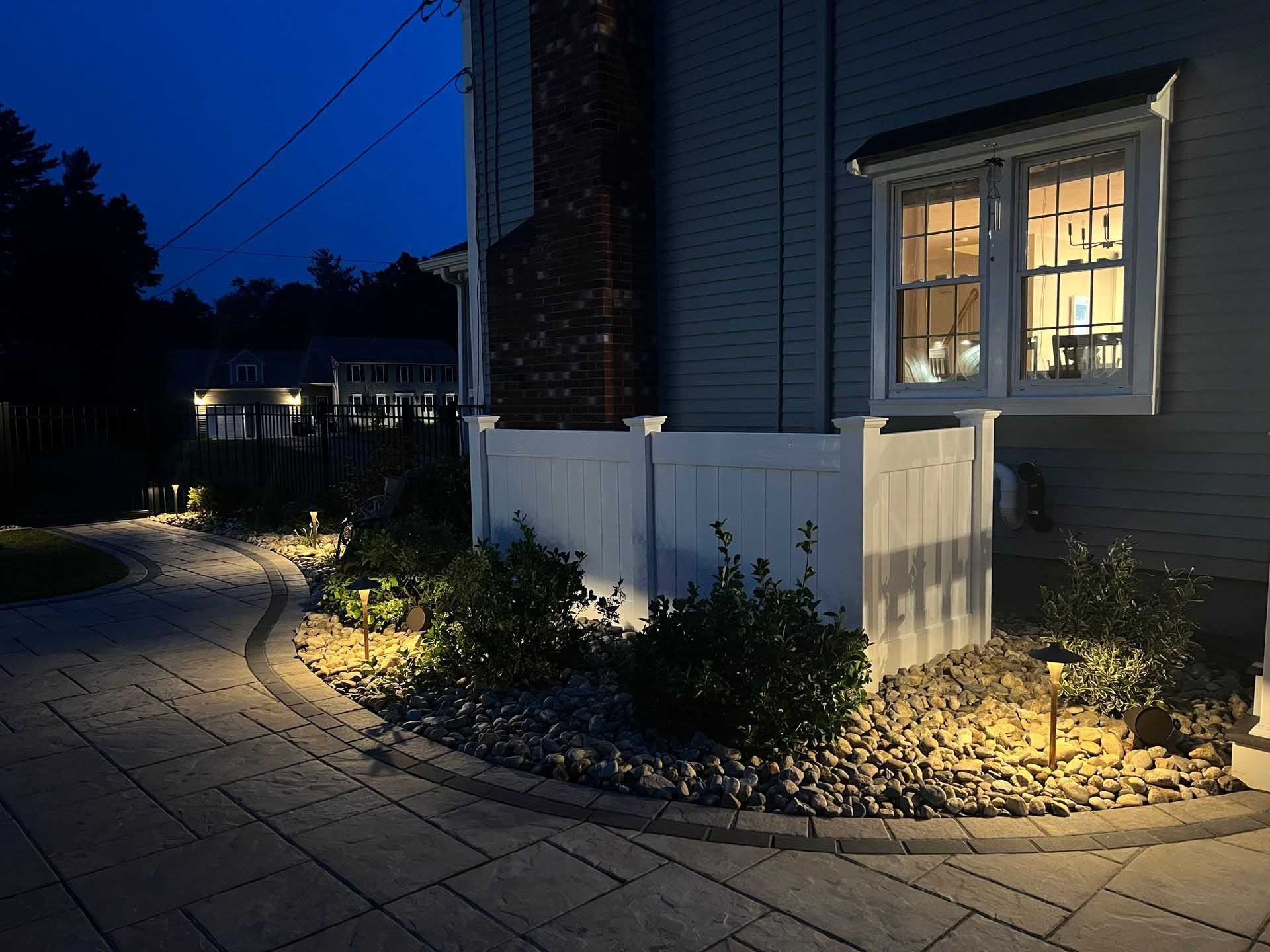Nighttime exterior shot of a house with illuminated landscaping and a lit window.