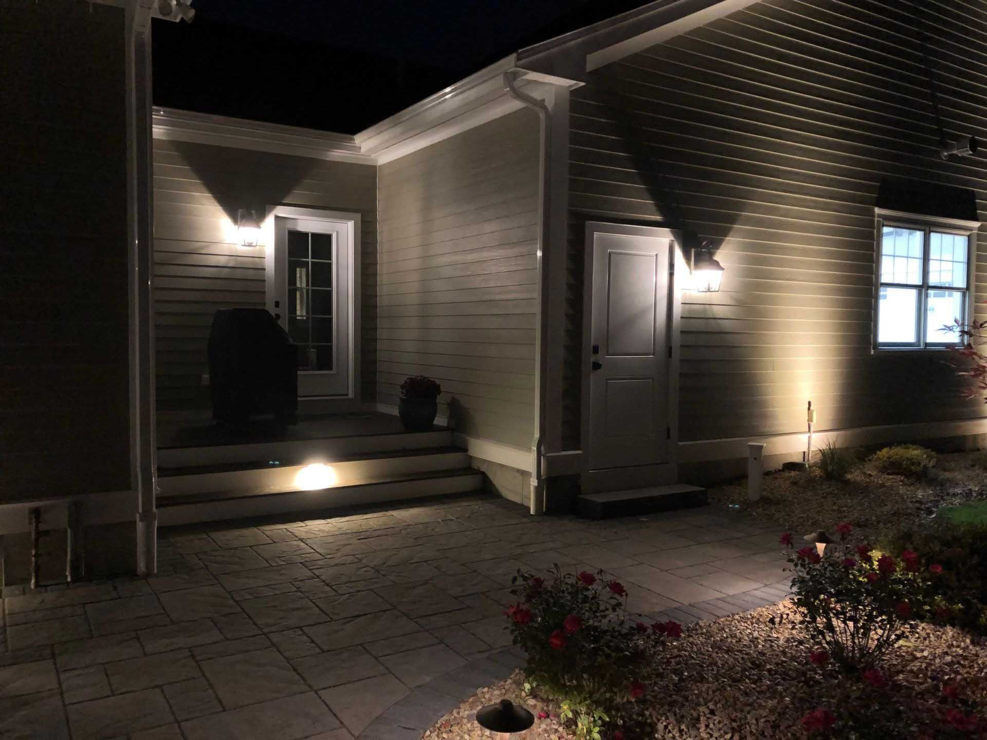 Nighttime view of a house with outdoor lighting illuminating the facade, doors, steps, and landscaping.