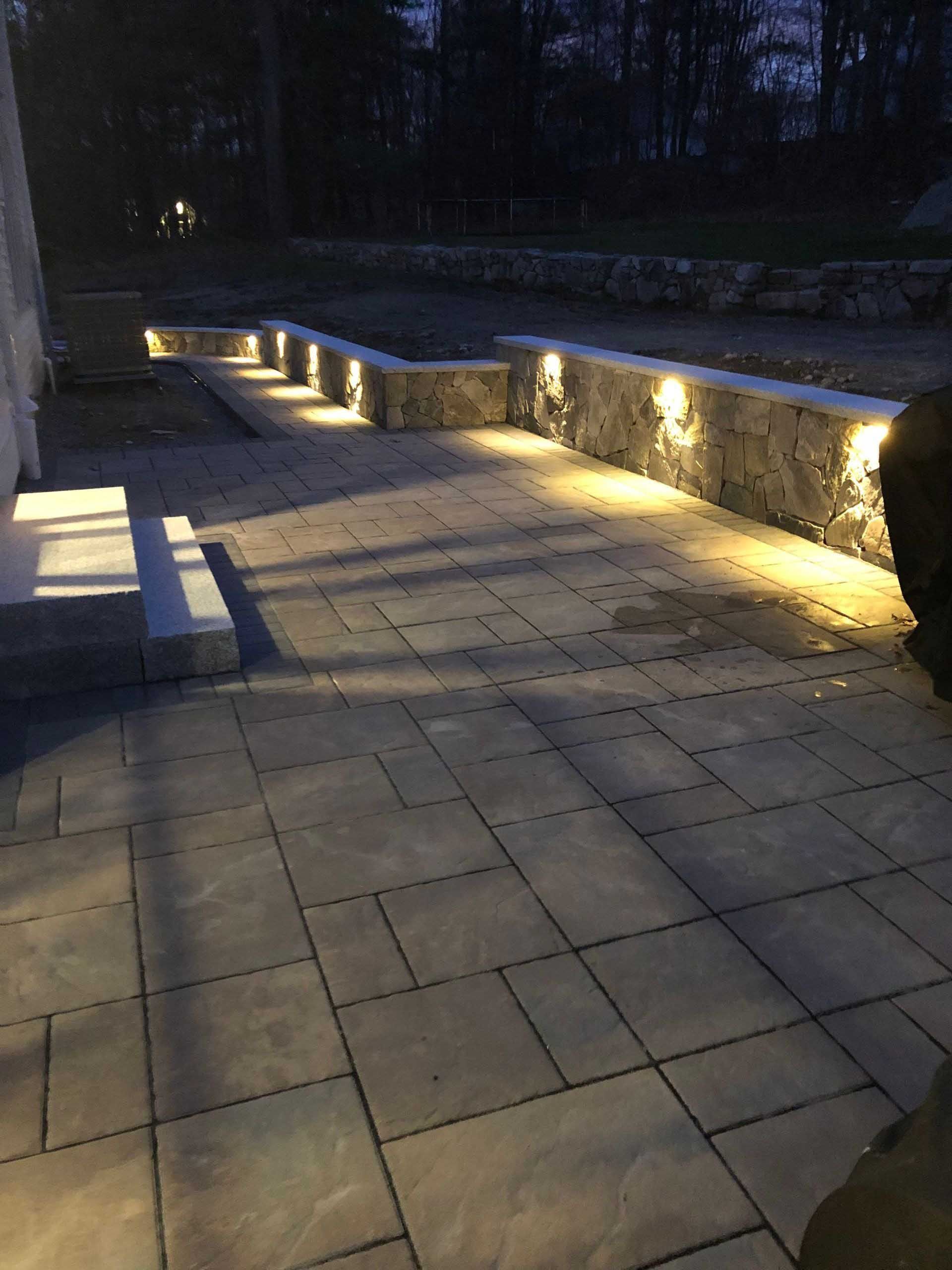 Stone patio with lit wall lights, steps, and a retaining wall at dusk.