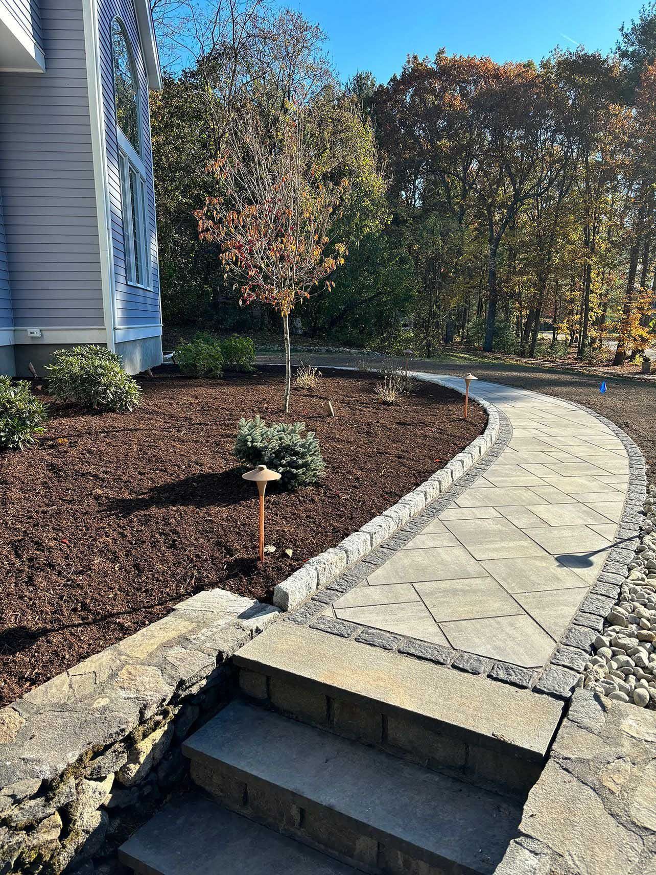 Stone path curves through a landscaped yard with a tree and stairs leading up to a house.