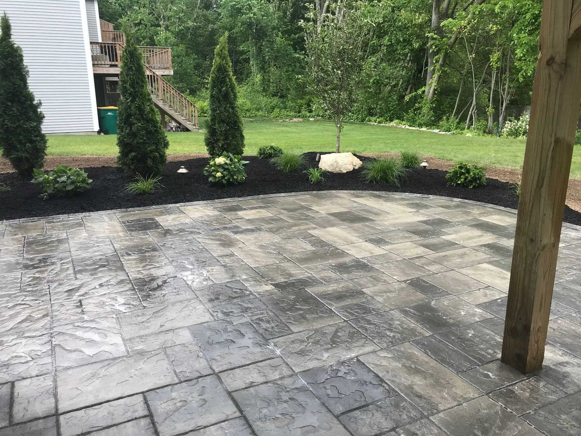 Stone patio with a flowerbed and evergreen trees; green grass and trees in background.