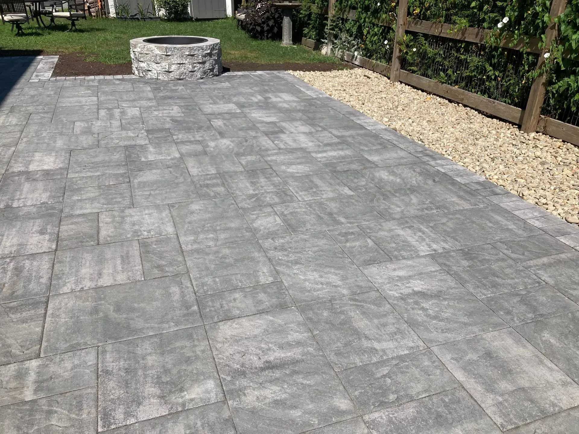 Gray stone patio with a fire pit, next to gravel and a wooden fence.