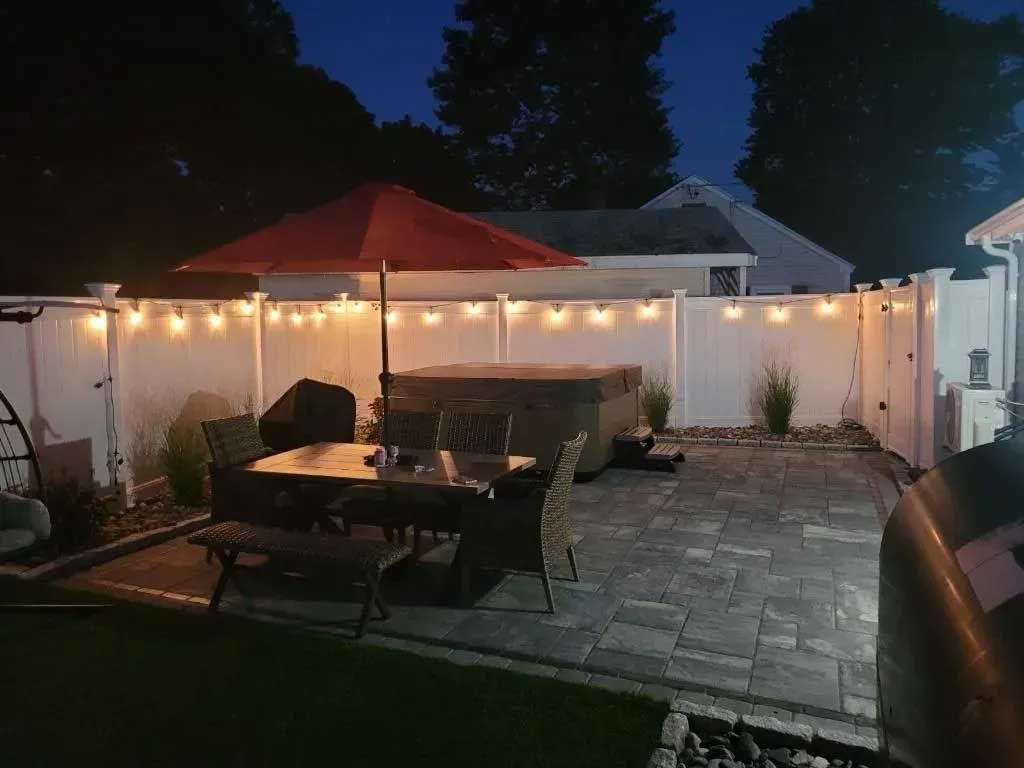 Backyard patio with dining set, hot tub, and string lights along a white fence.