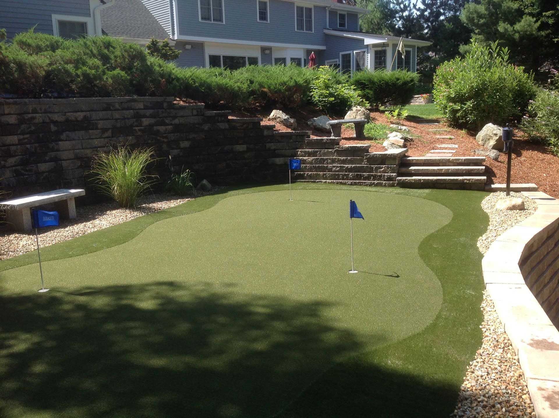 Backyard putting green with blue flag, stone steps, and landscaping, next to a two-story house.