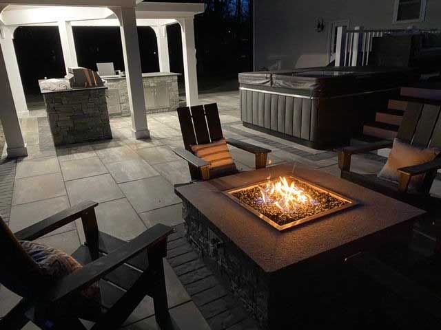 Patio with a lit fire pit, chairs, and a hot tub at night.