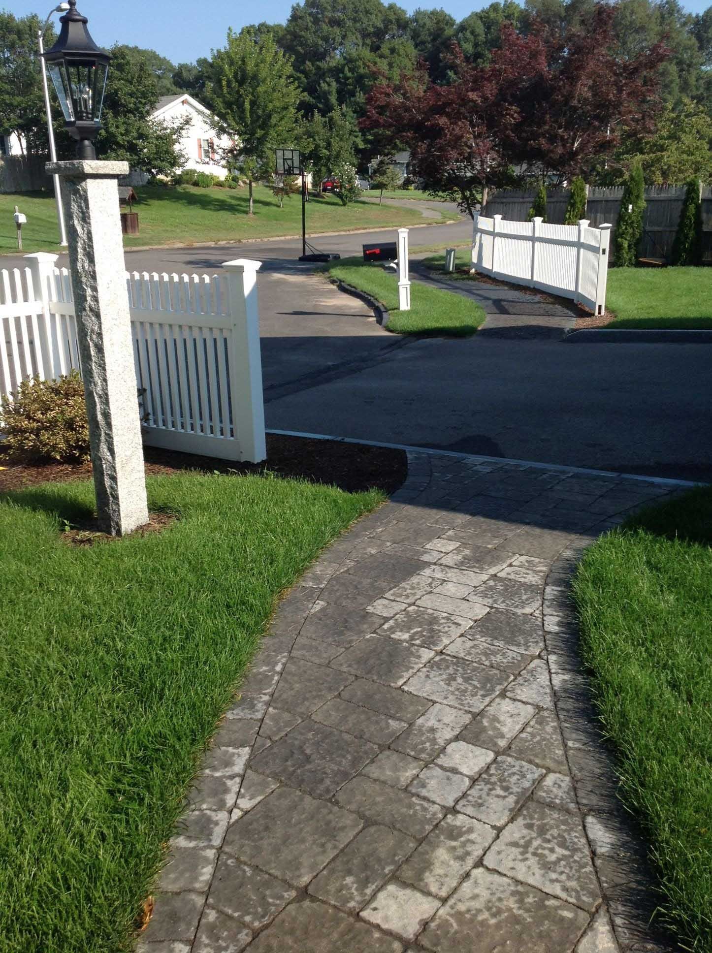 Brick walkway leads to white fence, opening onto a residential street with houses and trees.