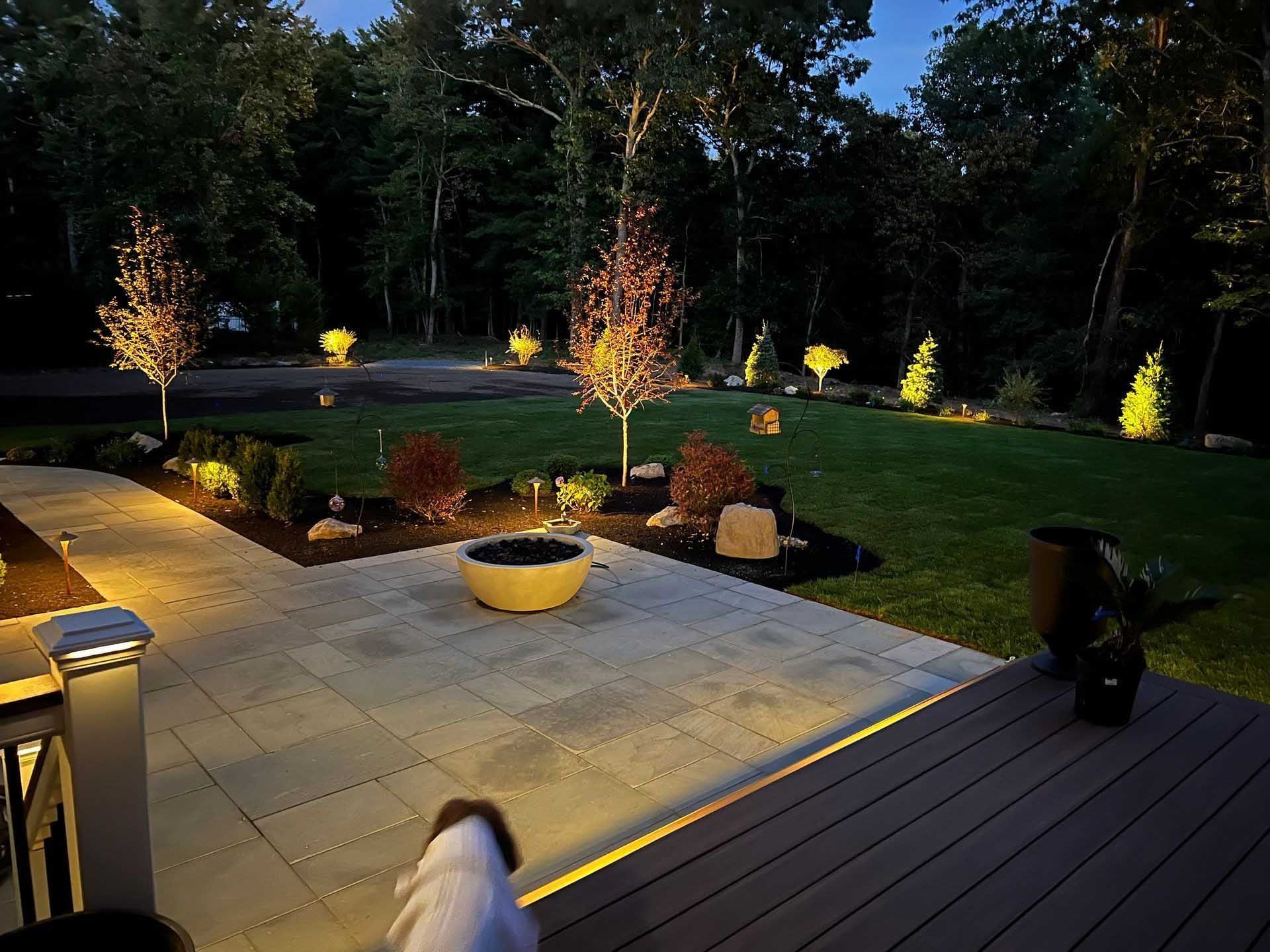 Nighttime outdoor scene: illuminated patio with landscaping, trees, and lights in a backyard.