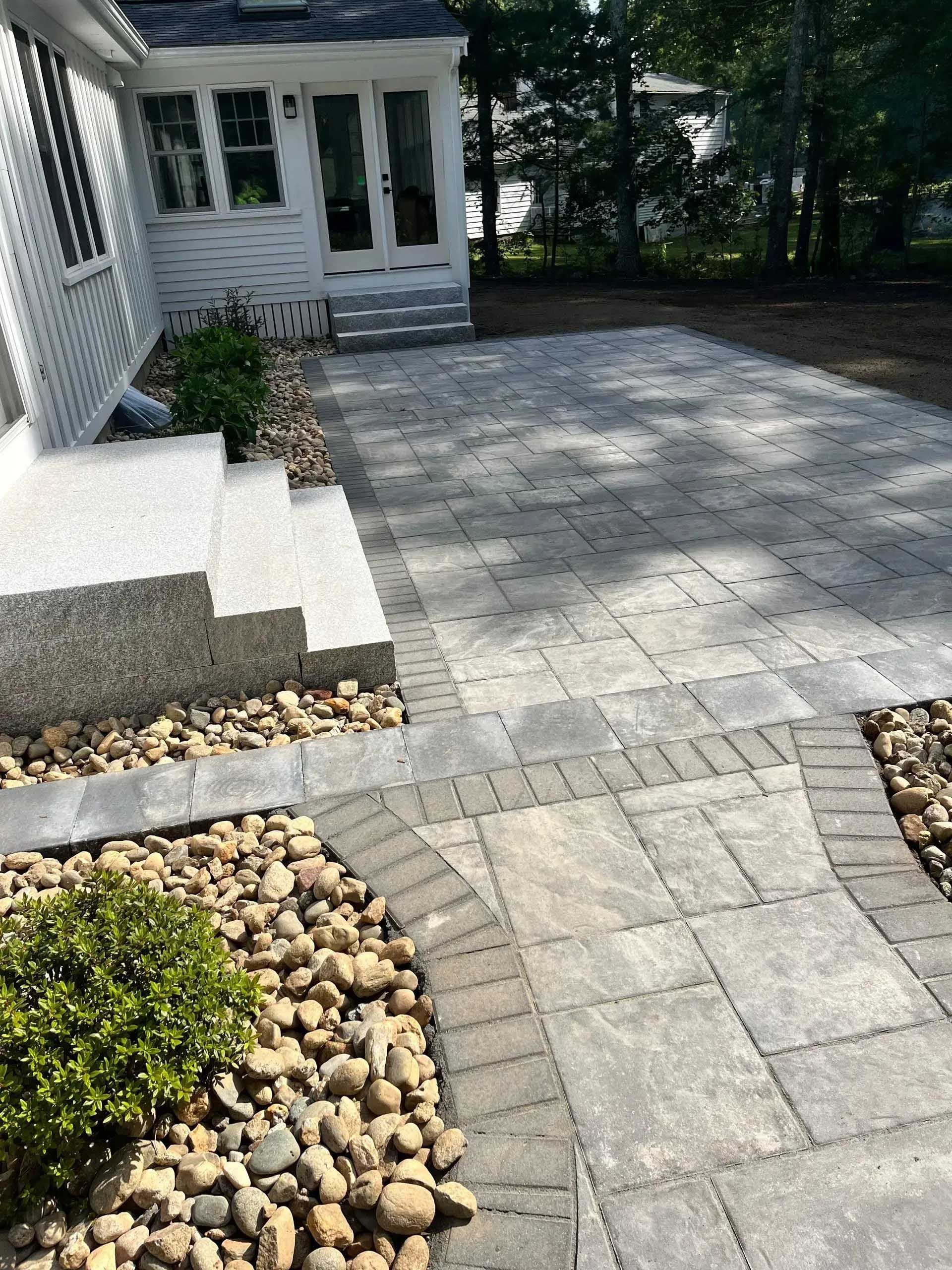 Stone patio with steps, pathway, and landscaping next to a white house.