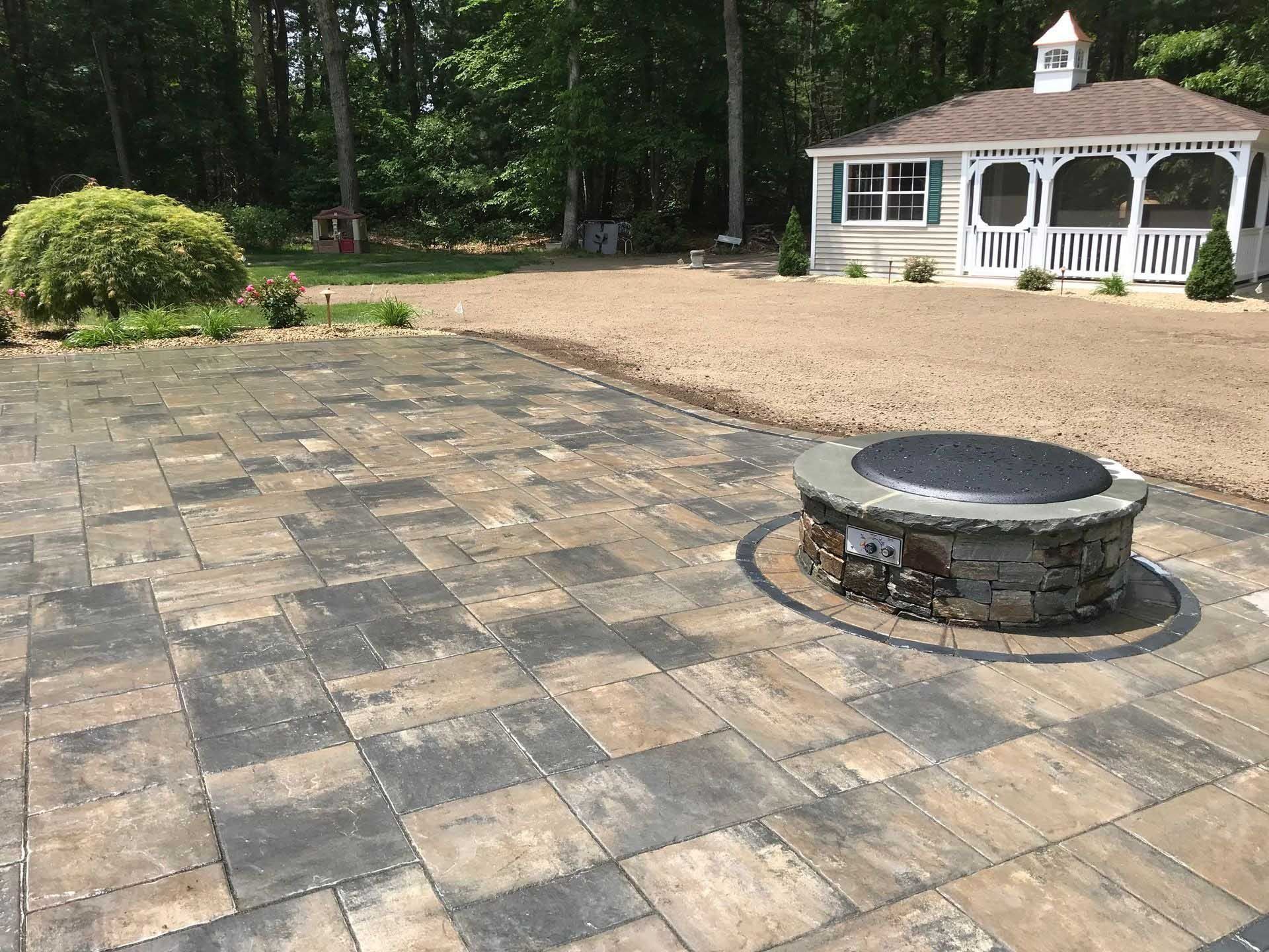 Stone patio with a fire pit, leading to a gazebo. Surrounding is gravel and trees.